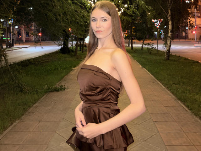 CaritaShover with long brown hair, strapless brown dress, holding it up, showing her tits, standing on a tiled pathway, trees and city lights in the background at night.