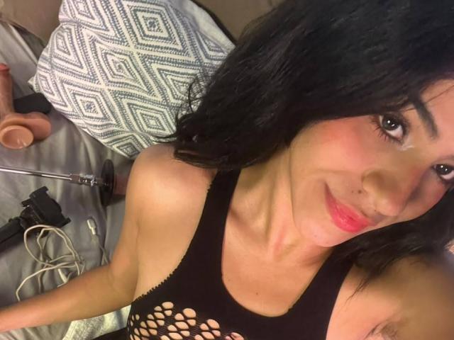 EvaGate with black hair, wearing a fishnet top, smiling at the camera, holding a dildo and a camera on a bed with a patterned pillow.