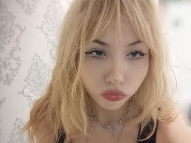 Eilene Szala, met blond haar, volle lippen en zwarte eyeliner, draagt een zilveren ketting en kijkt recht in de camera met een neutrale uitdrukking, waarbij haar kleine, ronde borsten zichtbaar zijn..