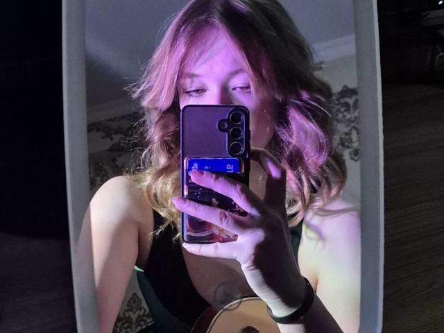 KimbraRenuart with pinkish-blonde wavy hair, black tank top, holding phone in mirror selfie, showing her tits and ass in reflection, wearing white wristband, in dimly lit room.