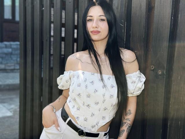Dindinlir-hot with long black hair, off-shoulder white floral top, and white pants, standing against a wooden fence, hands in pockets, tattoos on arms, and a black belt.