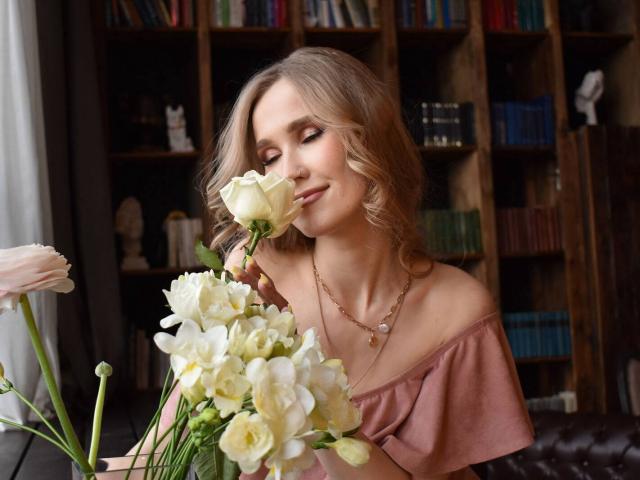 VeronikaWasher with wavy blonde hair, pink off-shoulder top, holding white flowers to her nose, wearing a necklace, in a cozy room with bookshelves in the background.