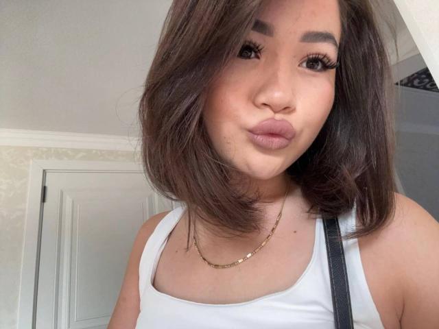 BarrieShelkoff has brown shoulder-length hair, wearing a white tank top, gold necklace, and pouty lips, with long eyelashes and a subtle makeup look, showing a cute expression.