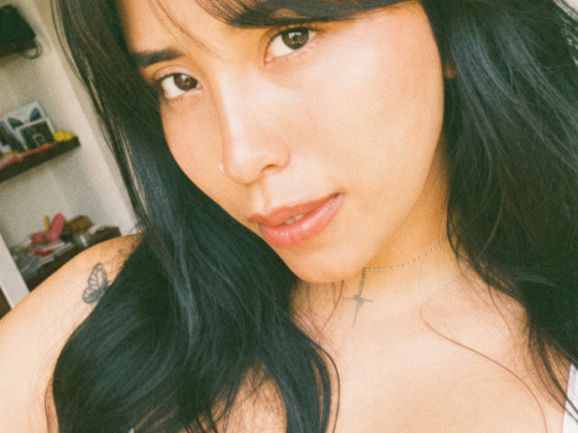 ElizabethMacy with long black hair, nose ring, and subtle makeup, showing her face and slightly parted lips, tattoo of a butterfly on her left shoulder, and a cross necklace.