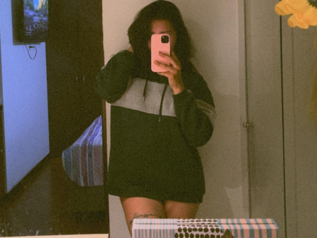 ElizabethMacy with dark curly hair, wearing a black and gray hoodie, taking a mirror selfie, showing her bare thighs and a tattoo on her left leg, holding a pink phone.