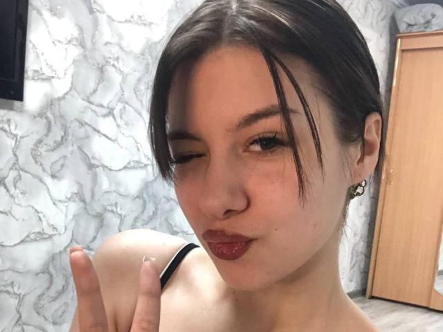 EchoKitanik with brown hair, winking, pouty red lips, black strap top, and diamond earrings, making a peace sign. Her tits are small and her skin is fair.