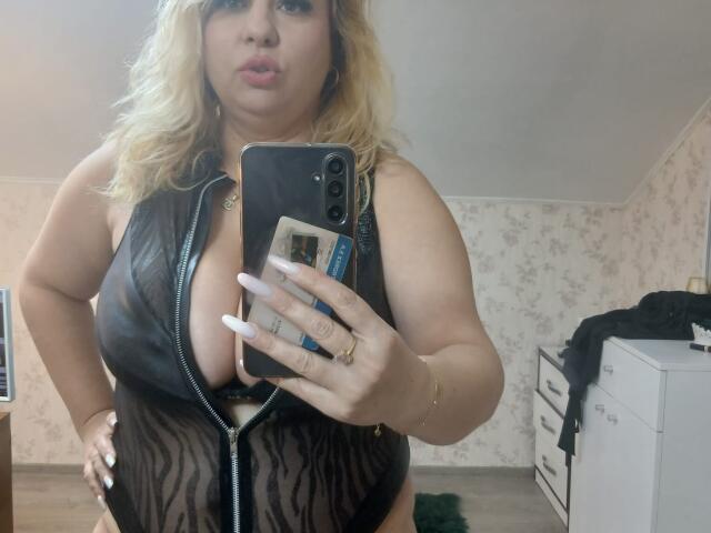 BelleInna mit blonden Haaren, voller Lippen, trägt einen schwarzen, durchsichtigen Overall mit Leopardenmuster und Reißverschluss, der ihre großen Brüste zeigt, und macht ein Selfie vor einem Spiegel mit ihrem Handy, in einem Schlafzimmer..