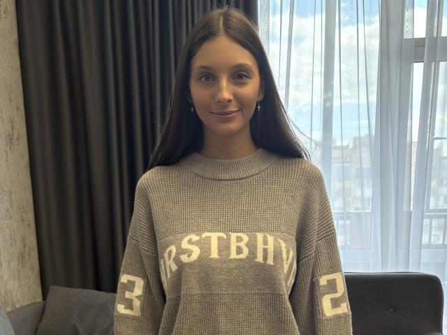Dottie Larson, avec de longs cheveux bruns, un pull gris avec l'inscription "RESTBHY" et des chiffres sur les manches, debout devant une fenêtre, souriant doucement, portant de petites boucles d'oreilles..