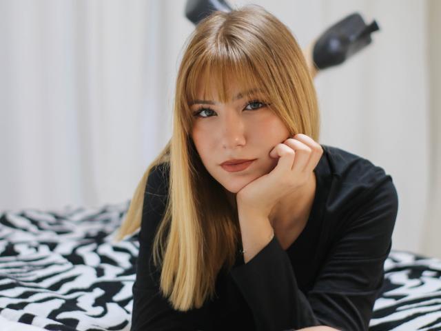 Chloe Kingsley, com cabelo loiro e franja, camisa preta de manga comprida, com o queixo apoiado na mão, deitada em uma cama com padrão de zebra, olhando para a câmera com uma expressão sedutora, maquiagem suave..