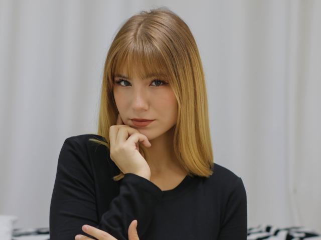 Chloe Kingsley, con cabello rubio hasta los hombros, una camisa negra de manga larga y una mano apoyada en la barbilla, mirando ligeramente hacia un lado con una sonrisa sutil. Sus senos son discretos..