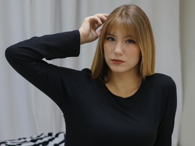 Chloe Kingsley mit glattem, schulterlangem braunem Haar, trägt ein schwarzes Langarmshirt, hat eine Hand im Haar, blickt in die Kamera, neutraler Gesichtsausdruck, kleine Brüste, blasse Haut, dezentes Make-up..