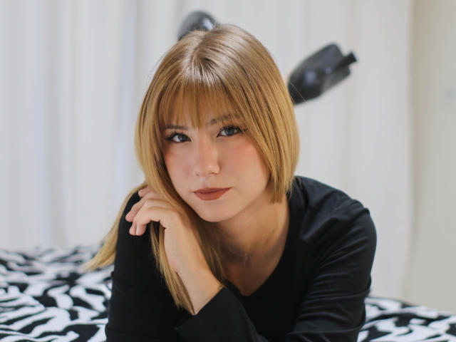 Chloe Kingsley, avec ses cheveux blonds, sa frange et son rouge à lèvres foncé, portant un haut noir, allongée sur un lit à motifs zébra, appuyant son menton sur sa main, regardant l'appareil photo..