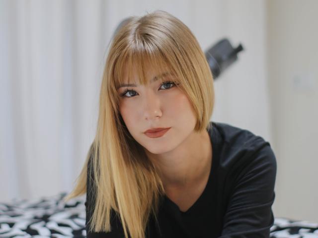 Chloe Kingsley, met blond haar, een zwart shirt en subtiele make-up, kijkt recht in de camera met een licht geopende mond. Haar borsten zijn klein en ze heeft een schattig gezicht..
