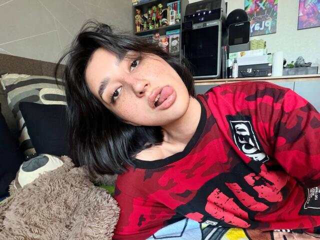 HeikeCarchi, with black hair and freckles, tongue out, wearing a red camo shirt, showing small tits, sitting on a bed with a fuzzy pillow.