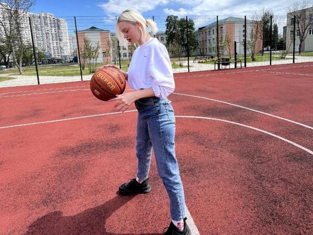 CrystalHope, blonde, white long-sleeve shirt, blue jeans, black sneakers, holding basketball on red outdoor court, standing, looking down, sunny day, blue sky, trees in background.