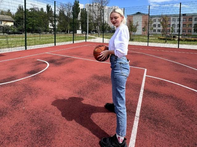 CrystalHope stands on a red basketball court, holding a basketball, wearing a white shirt, blue jeans, black shoes, with her shadow on the court.