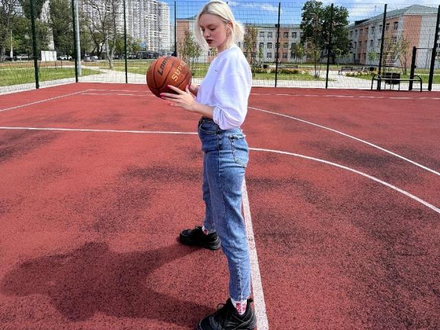 CrystalHope with blonde hair, white shirt, blue jeans, and black shoes, holding a basketball on a red outdoor court, standing with her back to the camera.