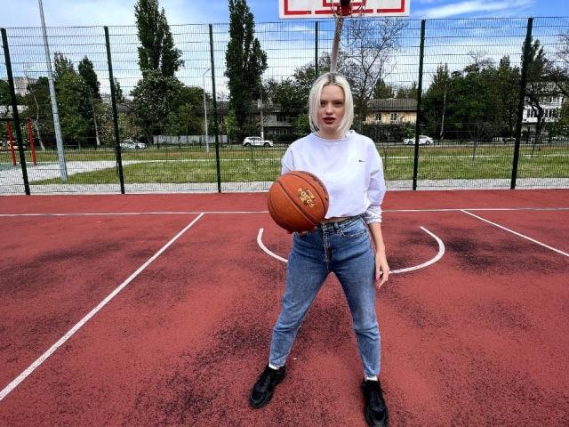 CrystalHope with blonde hair, wearing a white long-sleeve shirt, blue high-waisted jeans, black shoes, holding a basketball, standing on a red outdoor basketball court.