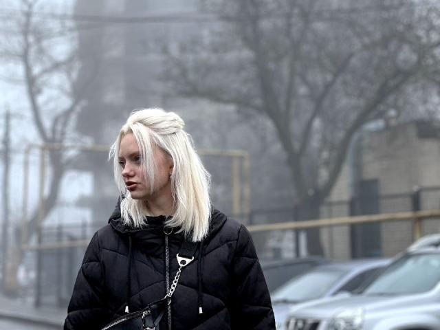 CrystalHope with pale blonde hair, black puffy jacket, chain purse, and serious expression, standing in foggy urban setting, cars and trees blurred in background.