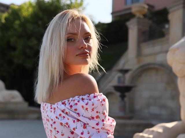 CrystalHope with blonde hair, off-shoulder floral dress, looking over her shoulder, sunlit, outdoor, stone fountain, soft smile, pretty, tits partially visible, background blurred.