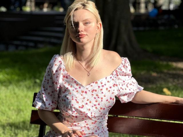 Blonde CrystalHope with small tits, wearing a white floral dress, sits on a wooden bench in sunlight, hand on her hip, looking slightly to the side, with a necklace.