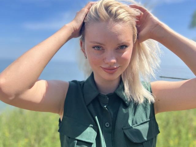 CrystalHope has blonde hair, blue eyes, and fair skin. She's wearing a green sleeveless shirt, standing with hands in her hair, smiling softly. She's got a cute ass.