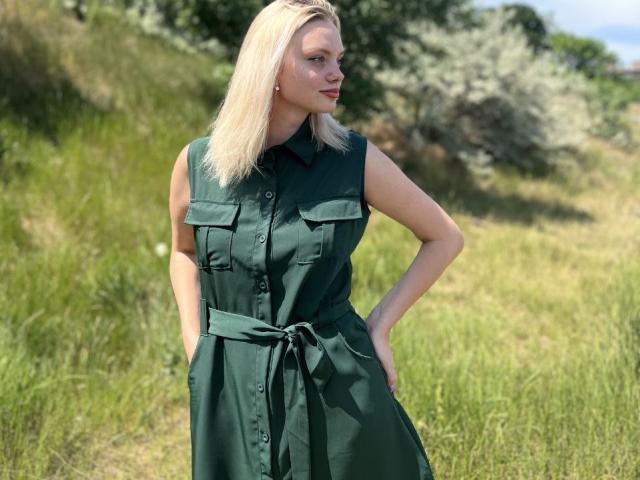 CrystalHope with blonde hair, green sleeveless dress with pockets, hands in pockets, standing in grassy field, looking to the side, sunny day, blue sky in background.