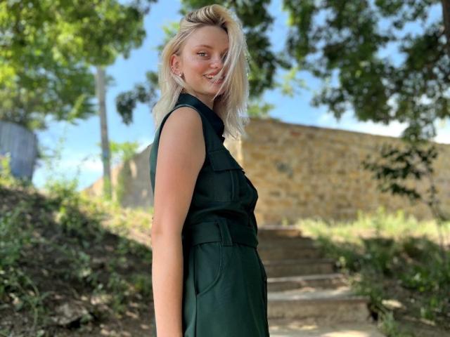 CrystalHope with blonde hair, smiling, wearing a sleeveless green dress, standing outdoors, her ass slightly visible, stone wall in background, green trees, clear blue sky, and sunny day.