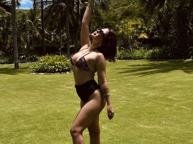 MedeaAllure tem um corpo curvilíneo e sensual, com seios grandes, vestindo um biquíni preto com estampa de leopardo, parte de baixo preta de cintura alta e óculos de sol, levantando um braço em uma área ensolarada e gramada..