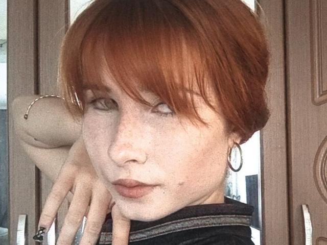 KasandraGrothe has red hair, freckles, and light skin. She's looking sideways with a serious expression, wearing a black top and hoop earrings. Her right hand touches her chin.