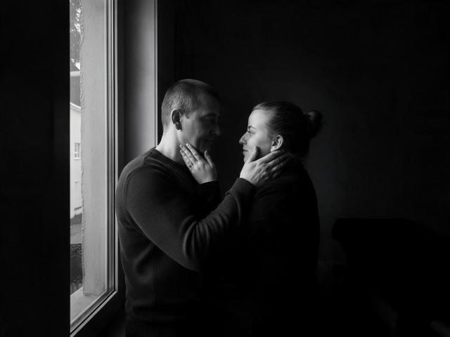 Black-and-white photo of AlfixKellys with short hair and AlfixKellys with a bun, standing by a window, touching faces, smiling, wearing dark long-sleeve shirts, in soft light.