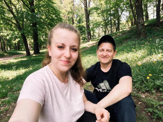 Two AlfixKellys sitting in a forest, one with brown hair in a pink shirt, the other with black hair in a black Adidas shirt, both smiling.