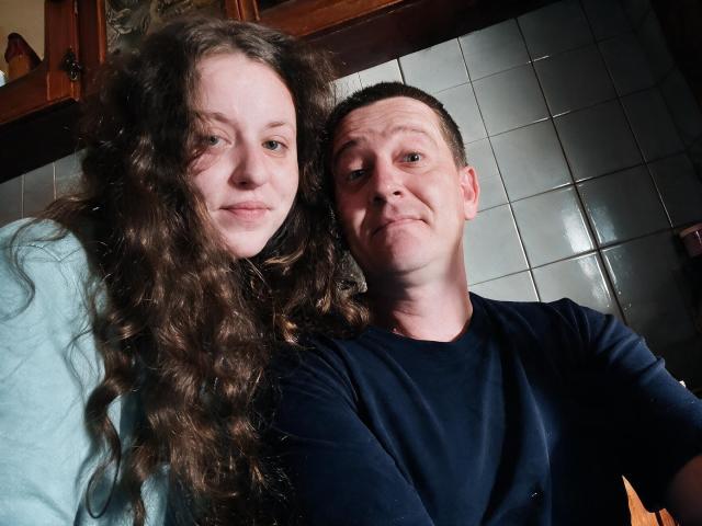 AlfixKellys with long curly brown hair and pale skin, wearing a light blue shirt, stands next to AlfixKellys with short brown hair and pale skin, wearing a navy shirt.