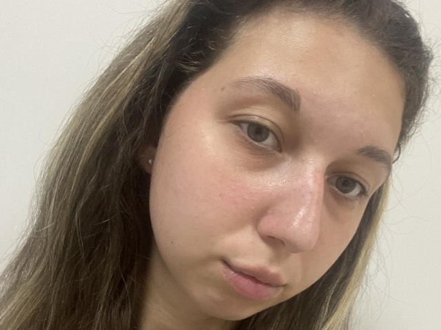 Sanella's face, showing light skin, brown eyes, and long brown hair with blonde highlights, has a slight smile and subtle pinkish nose with a small blemish.