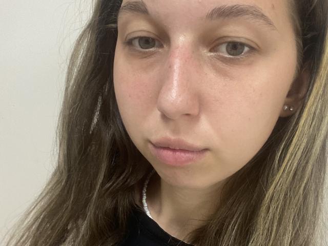 Sanella's face, showing pale skin, brown eyes, and slightly parted pinkish lips. She has long brown hair with blonde highlights, and a small silver earring on the right.