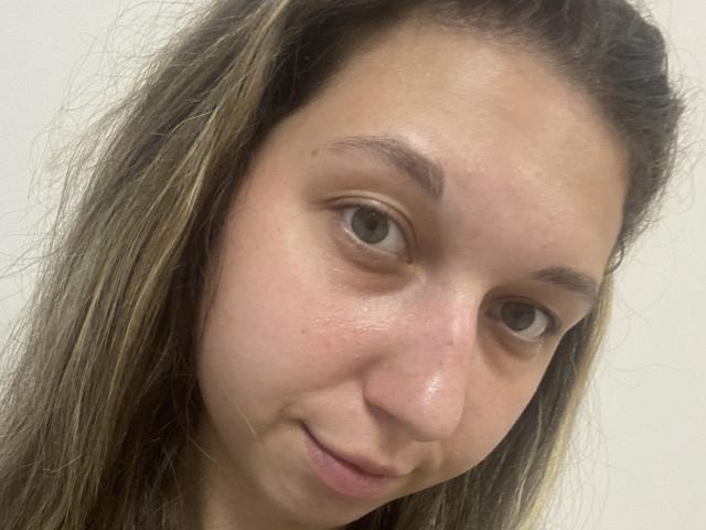 Sanella's face, showing brown eyes, slightly flushed skin, and a small smile. She has long, messy brown hair with blonde highlights. Her nose is small and she looks cute.