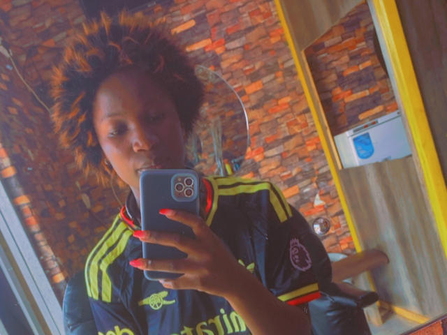JazzBerry met donkerbruine huid, krullend oranje haar, gekleed in een zwarte sportjas met gele strepen, die een selfie maakt met een blauwe telefoon, met rode nagels..