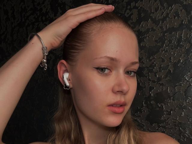 LillianaWhang with blonde hair, green eyes, and pink lips, wearing white earbuds, a silver bracelet, and hand on head, in a dark textured background.