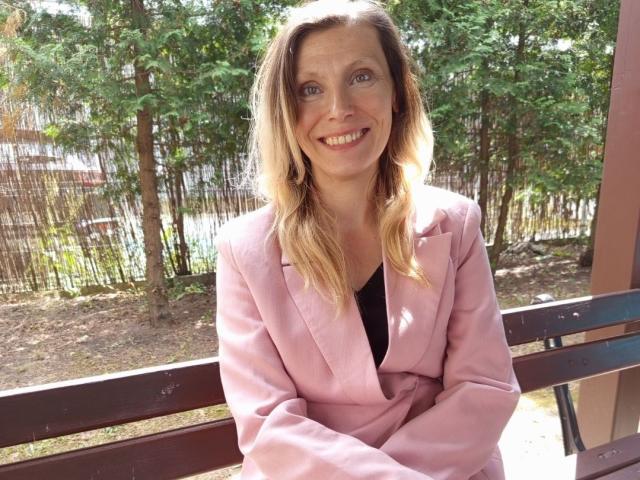Catherine, con capelli biondi, un blazer rosa, una maglietta nera, sorridente, con le braccia incrociate, seduta su una panchina di legno, alberi verdi sullo sfondo, luce solare, rilassata, attraente, occhi azzurri, corporatura media..