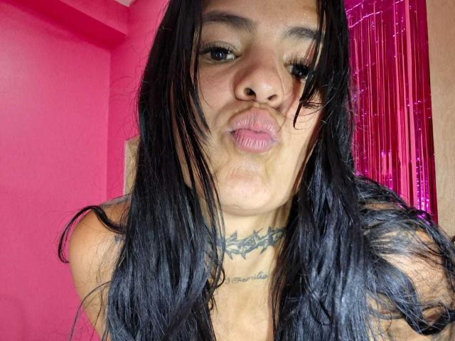 InkSinner69, con cabello negro, frunciendo los labios, mostrando un cuello tatuado, sin ropa, frente a una pared rosa con hilos morados. Sus pechos no son visibles..
