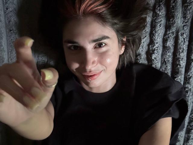 TameikaMacclellan, com unhas amareladas, sorrindo, deitada em uma superfície cinza texturizada, vestindo uma camisa preta e apontando para a câmera com os lábios vermelhos ligeiramente entreabertos..