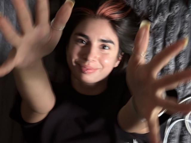 TameikaMacclellan, com as mãos estendidas, sorrindo com um leve franzir de lábios, olhos castanhos, cabelo escuro com pontas rosa, vestindo uma camisa preta, deitada, com os dedos espalhados..