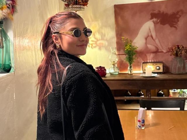 BiancaRey con i capelli tinti di rosa, indossa un cappotto nero e peloso, porta occhiali da sole rotondi, è in piedi in una stanza accogliente, guarda indietro per le spalle, sorridendo..