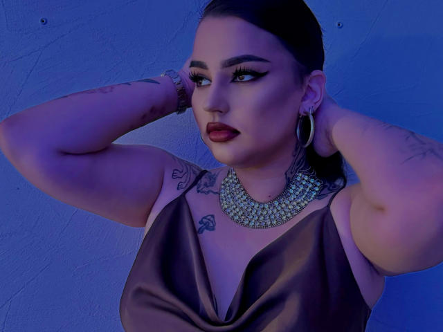 NikkiFley, avec des tatouages, du rouge à lèvres foncé et des boucles d'oreilles pendantes, portant une robe en satin marron, les mains derrière la tête, éclairage bleu, regardant sur le côté, un grand collier orné de bijoux..