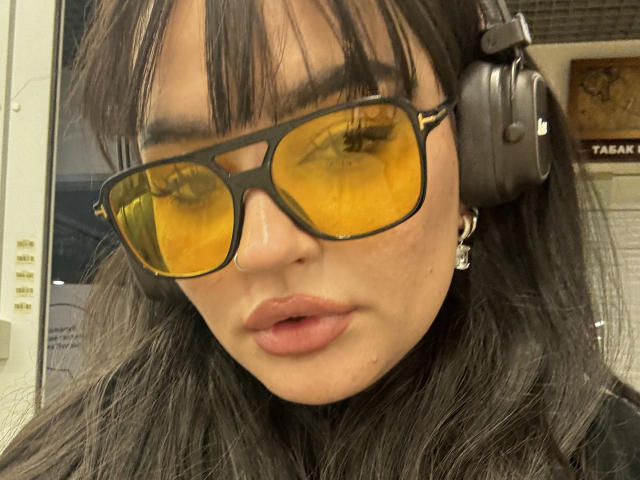 NikkiFley, con occhiali da sole di colore giallo, cuffie nere e labbra leggermente dischiuse, ha capelli neri e lisci, e un piercing al naso, appare seria in un selfie..