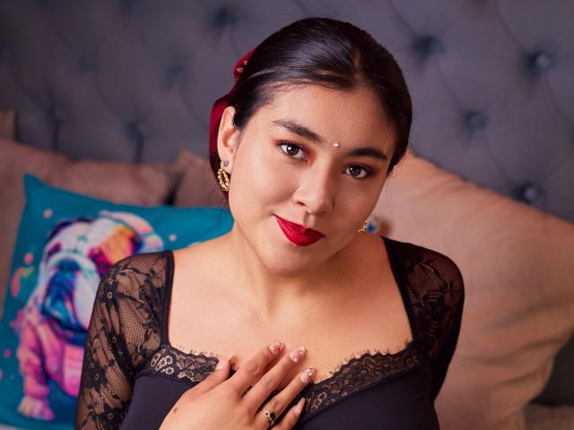HannaGoez con capelli scuri, rossetto rosso, top in pizzo nero, orecchini dorati, punto sulla fronte e mano sul petto, che guarda la fotocamera, sorridendo, carina, sexy, latina, seno di media grandezza..
