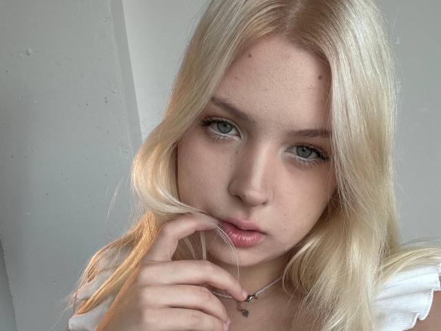 BelleGagnier with blonde hair, green eyes, and fair skin, touching her lips with a finger, wearing a white shirt and silver necklace, looking slightly pouty.