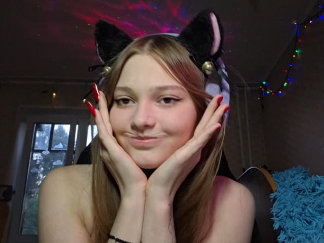BrittanieManino with cat ears, red nails, and hands on cheeks, smiling, has long blonde hair, and is topless, showing her tits, in a room with colorful lights.