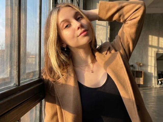 Bethany Aceves, avec ses cheveux blonds, portant un manteau beige par-dessus un débardeur noir, se tient près d'une fenêtre, la lumière du soleil projetant des rayures sur son visage, une main derrière la tête, un collier..