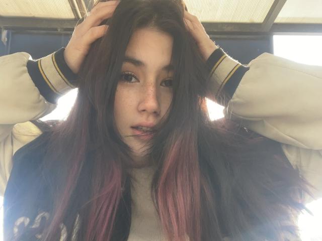 PattiTropea with long dark hair, hands in her hair, wearing a white and blue varsity jacket, looking down with freckles and a slight pout, sunlight in the background.